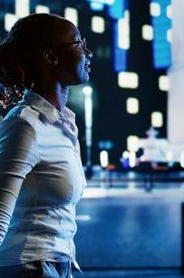 微笑的非裔美国妇女在夜晚漫步在城市的林荫大道上, pointing at interesting billboards on skyscrapers. 在灯火照亮的街道上漫步的女商人